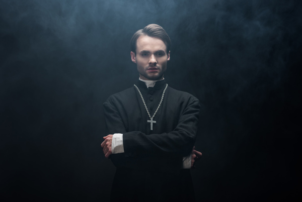 молодий серйозний католицький священик, що стоїть з схрещеними руками на чорному тлі з димом
 - Фото, зображення
