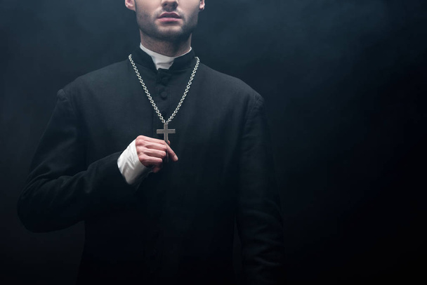 cropped view of catholic priest touching silver cross on his necklace on black background with smoke - Foto, Imagen