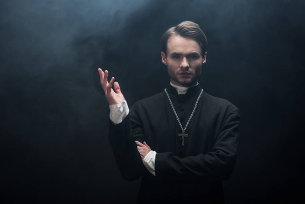 молодий впевнений католицький священик, що стоїть з відкритою рукою на чорному тлі з димом
 - Фото, зображення