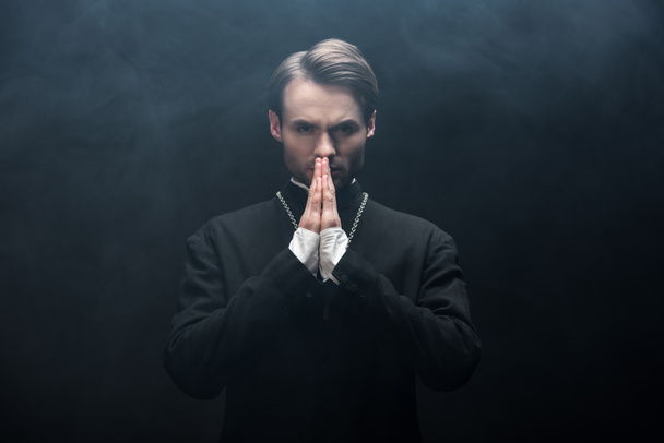 serious catholic priest looking at camera while praying on black background with smoke - Photo, Image