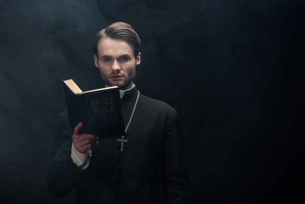 young serious catholic priest looking at camera while reading bible on black background with smoke - Φωτογραφία, εικόνα
