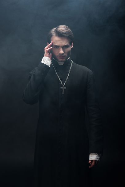 young thoughtful catholic priest touching head while looking at camera on black background with smoke - Foto, imagen