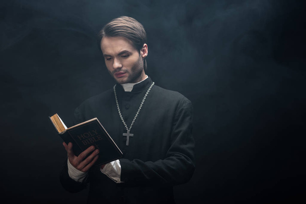attentive catholic priest reading holy bible on black background with smoke - Photo, Image