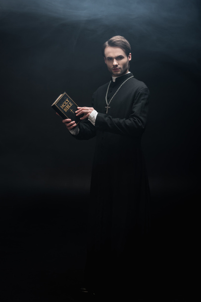 volledige weergave van strikte katholieke priester houden heilige bijbel en kijken naar camera op zwarte achtergrond met rook - Foto, afbeelding