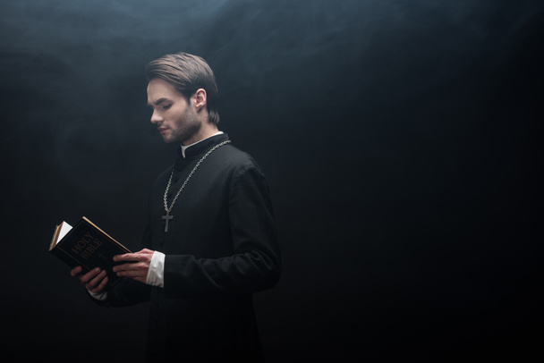 concentrated catholic priest reading holy bible on black background with smoke - Фото, зображення