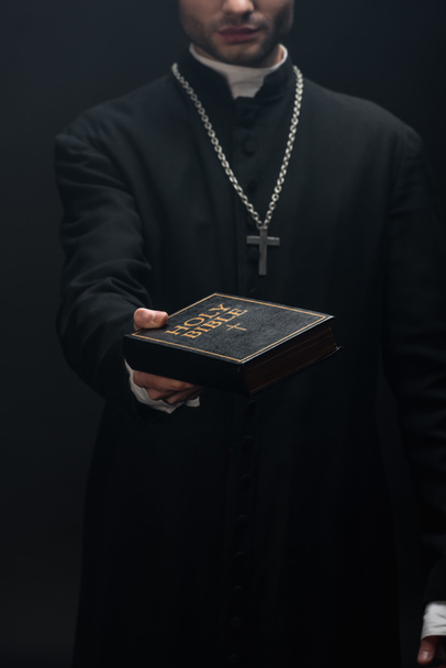 visão cortada de padre católico segurando Bíblia sagrada em mão estendida isolado em preto
 - Foto, Imagem