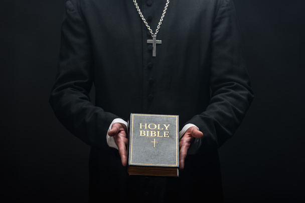 cropped view of catholic priest holding holy bible isolated on black - Foto, Imagem