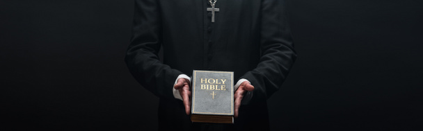vista cortada de padre católico segurando bíblia sagrada isolado em preto, tiro panorâmico
 - Foto, Imagem
