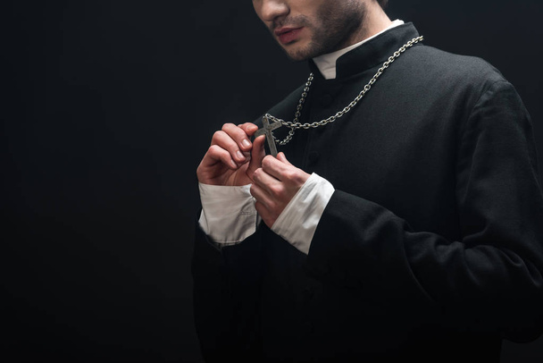 cropped view of catholic priest looking at silver cross on his necklace isolated on black - Photo, Image