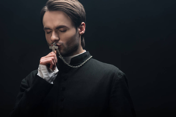 молодой вдумчивый католический священник целует серебряный крест с закрытыми глазами, изолированными на черном
 - Фото, изображение