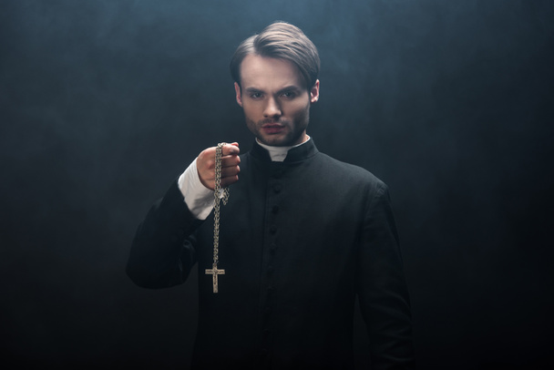 confident catholic priest holding necklace with cross while looking at camera on black background with smoke - Fotoğraf, Görsel