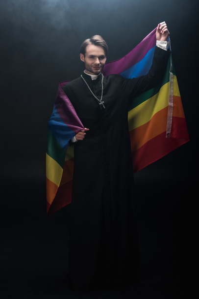 full length view of smiling catholic priest holding lgbt flag while looking at camera on black background with smoke - Foto, afbeelding