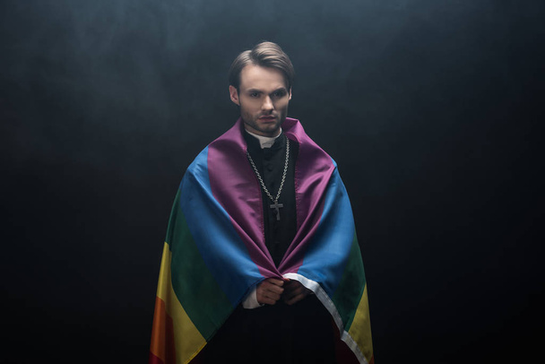 serious catholic priest wrapped in lgbt flag while looking at camera on black background with smoke - Photo, Image