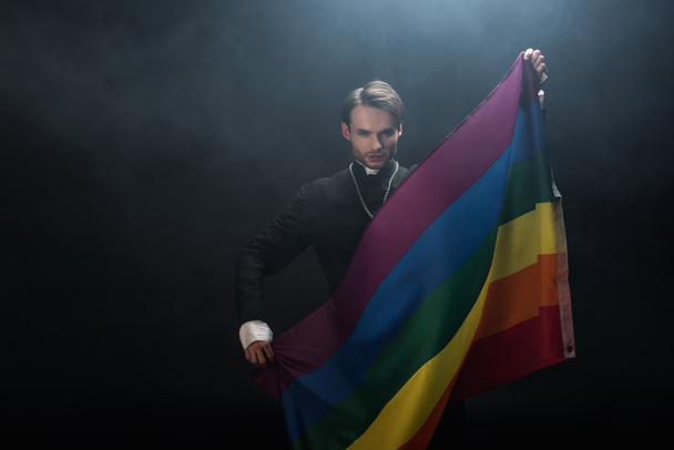 confident catholic priest holding lgbt flag while looking at camera on black background with smoke - Photo, Image