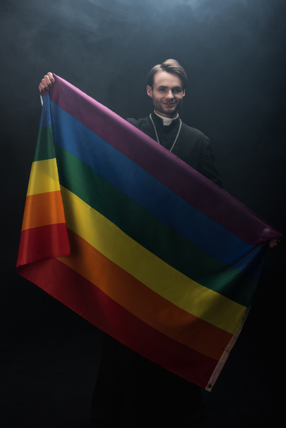 sonriente sacerdote católico sosteniendo la bandera lgbt mientras mira a la cámara sobre fondo negro con humo
 - Foto, Imagen