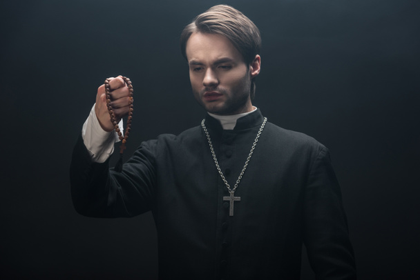 young tense catholic priest looking at wooden rosary beads isolated on black - Foto, Imagem