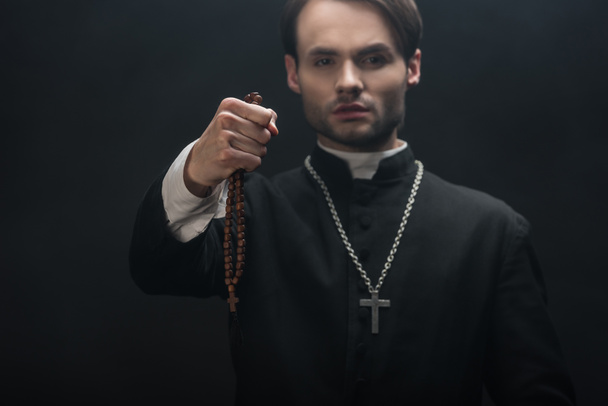 selective focus of young confident catholic priest holding wooded rosary beads in outstretched hand isolated on black - Фото, зображення