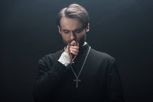вдумчивый католический священник целует серебряный крест с закрытыми глазами, изолированными на черном
 - Фото, изображение