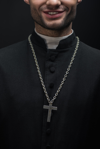 黒で隔絶されたネックレスの上に銀の十字架を持つ笑顔のカトリック聖職者のクロップドビュー - 写真・画像