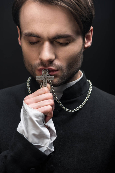 młody troskliwy katolicki ksiądz całuje krzyż z zamkniętymi oczami odizolowanymi na czarno - Zdjęcie, obraz