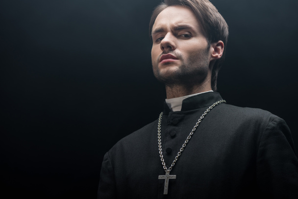low angle view of young tense catholic priest looking at camera isolated on black - Foto, imagen