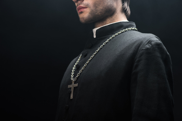 przycięty widok katolickiego księdza w czarnej sutannie ze srebrnym krzyżem na naszyjniku odizolowanym na czarno - Zdjęcie, obraz