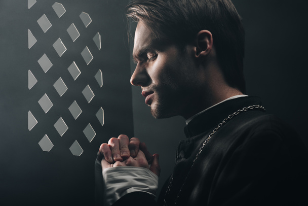 молодий вдумливий католицький священик молиться закритими очима в темряві біля конфесійної решітки з променями світла
 - Фото, зображення