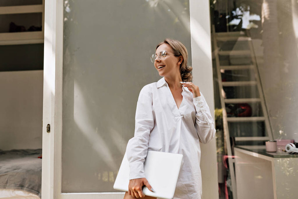 junge erfolgreiche Frau im weißen Hemd mit Laptop, die in der Morgensonne im Büro spaziert - Foto, Bild