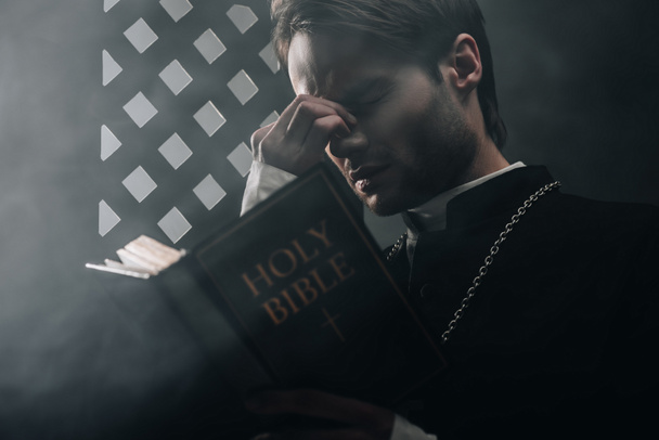 młody troskliwy katolicki ksiądz dotykający twarzy podczas czytania Biblii w pobliżu konfesjonalnej kratki w ciemności z promieniami światła - Zdjęcie, obraz