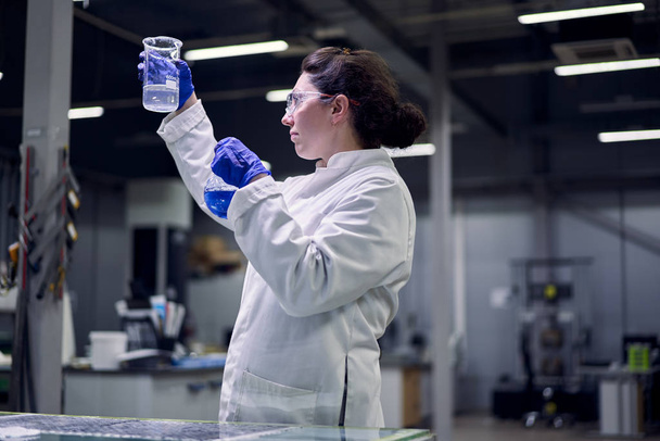 Brunette lab woman in glasses and white coat with flask with blue liquid in her hands conducts experiments - Фото, изображение