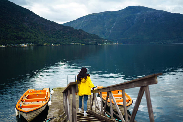 The girl tourist in a yellow jacket posing by the lake in Norway. Active woman relaxing and enjoys freedom near the boat by the lake against the backdrop of the mountains. Travelling, lifestyle. - Photo, image