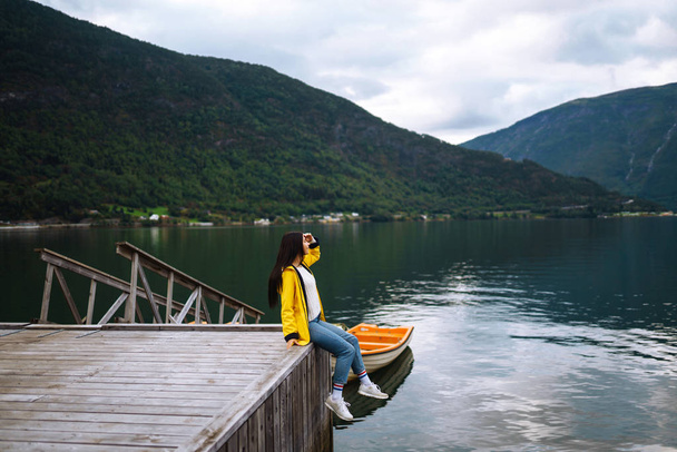 The girl tourist in a yellow jacket posing by the lake in Norway. Active woman relaxing and enjoys freedom near the boat by the lake against the backdrop of the mountains. Travelling, lifestyle. - Foto, Bild