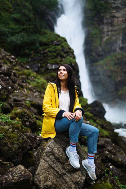 The girl tourist in a yellow jacket enjoys freedom against the backdrop of the waterfall. Young woman looking on the majestic waterfall in the Norway. Travelling, lifestyle, adventure, concept. - Foto, Bild