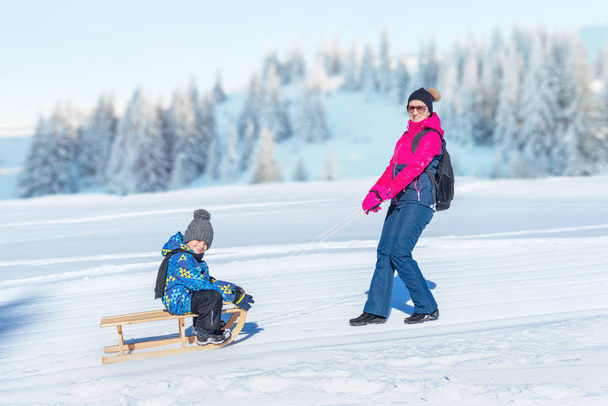 母は少年と一緒に雪そりをドラッグします。山を背景に。コンセプトodは冬の間子供を健康に保つ  - 写真・画像