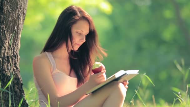 Girl sitting in park reading a book and eating an apple under tree - Záběry, video