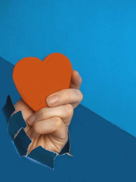 Kék szakadt papír lyuk, kéz gazdaság piros szív, szerelem, szenvedély szimbólum felett trendi kék színű háttér üzenet - Fotó, kép