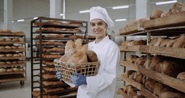 Bella signora panettiere in uniforme bianca in possesso di un cesto con pane fresco e sorridente carino davanti alla fotocamera
 - Foto, immagini