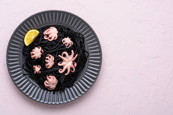 top view czarne spaghetti z atramentem cuttlefish z ośmiornicami i plasterkami cytryny w szarym talerzu na różowym tle, koncepcja kuchni śródziemnomorskiej - Zdjęcie, obraz