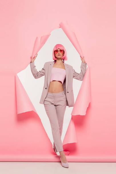 stylish girl in trendy suit, sunglasses and pink wig posing in torn paper, on white - Foto, imagen