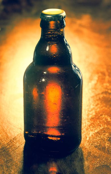 Unopened unlabeled brown glass bottle of beer backlit on a wooden surface with corner vignetting for your advertising or marketing - Foto, Imagen