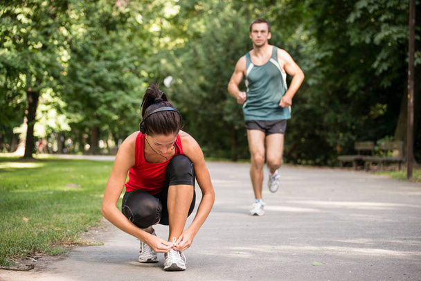 Young sport woman tying shoelace, man in behind is jogging - Fotoğraf, Görsel