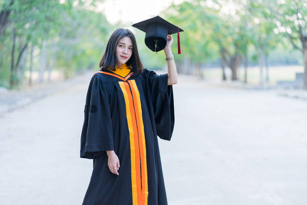 Portrait of a young cheerful female graduate wearing academic gown holding graduate cap celebrates her university degree in commence day in the college campus. - Photo, Image