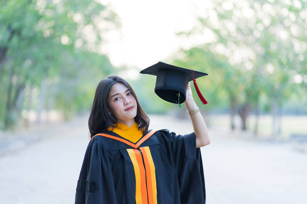 Portrait of a young cheerful female graduate wearing academic gown holding graduate cap celebrates her university degree in commence day in the college campus. - Photo, Image