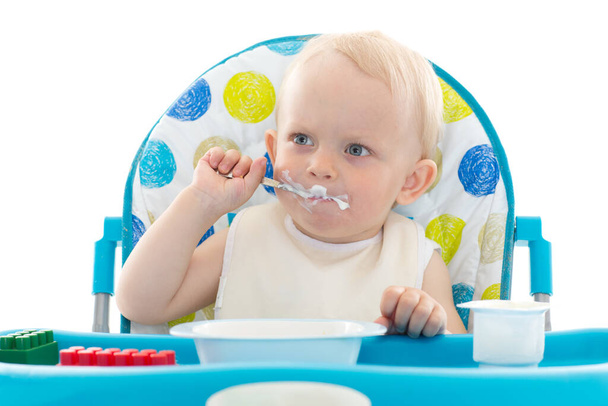 Sweet baby learning to eat with spoon sits on baby chair on a white background. - Photo, image