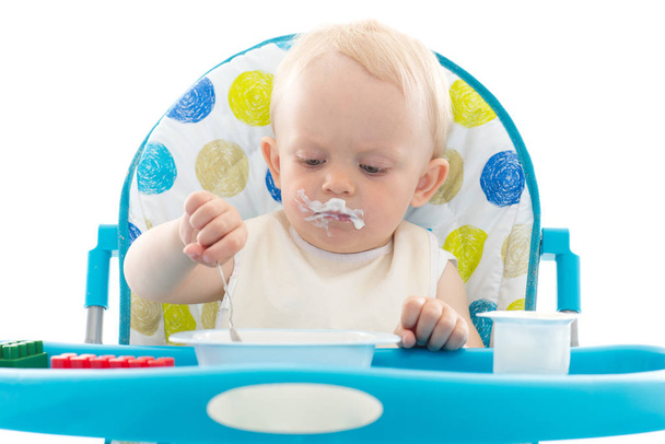 Sweet baby learning to eat with spoon sits on baby chair on a white background. - Zdjęcie, obraz