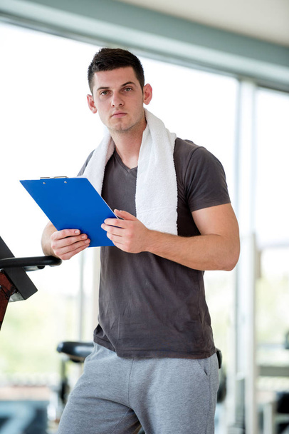 Portrait of a smiling male trainer with clipboard standing in a bright gym - Foto, afbeelding