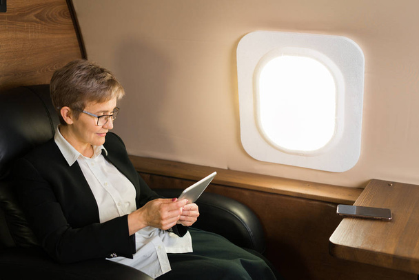 beautiful aged woman in the cabin of a private plane in a chair with a tablet in her hands - Фото, изображение