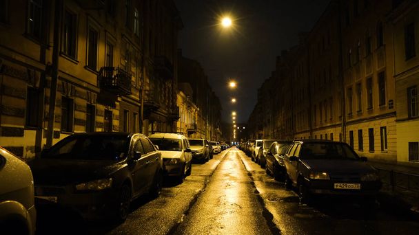 European street in light of yellow lanterns at night with cars in Parking. Concept. Straight beautiful street with residential buildings and parked cars lit by yellow lanterns at night - 写真・画像