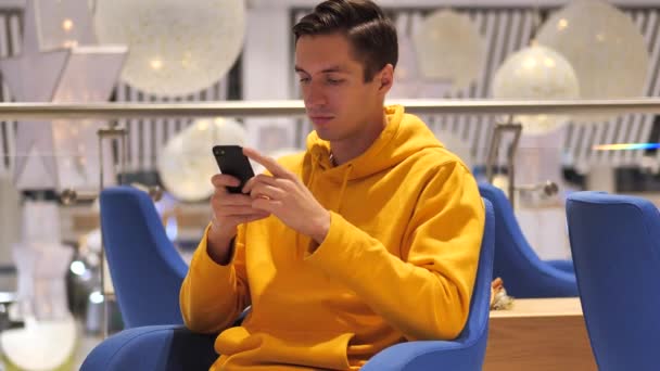 Young man in a yellow hoodie sits in a cafe mall shopping center and uses a modern smartphone - Séquence, vidéo
