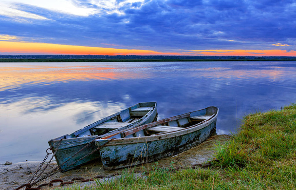 Due vecchie barche blu-verdi ormeggiate da una catena metallica sulla riva di un fiume calmo sullo sfondo del luminoso sole nascente
. - Foto, immagini